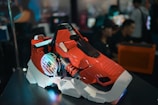 A futuristic, red and white gaming PC case with colorful LED lights and industrial design elements is displayed prominently. The background shows blurred, out-of-focus people and computer setups, suggesting a tech expo or gaming event.