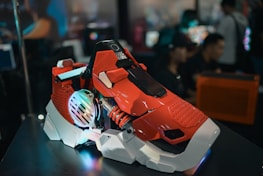A futuristic, red and white gaming PC case with colorful LED lights and industrial design elements is displayed prominently. The background shows blurred, out-of-focus people and computer setups, suggesting a tech expo or gaming event.