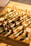 A well-lit chessboard with a full set of chess pieces arranged for the start of a game, casting long shadows across the board. The chess pieces are made of wood, and the board features alternating light and dark squares. The sunlight creates a warm and inviting atmosphere.