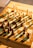 A well-lit chessboard with a full set of chess pieces arranged for the start of a game, casting long shadows across the board. The chess pieces are made of wood, and the board features alternating light and dark squares. The sunlight creates a warm and inviting atmosphere.