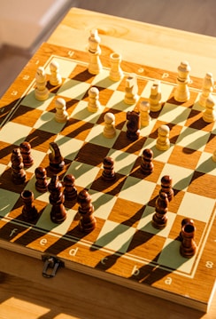A well-lit chessboard with a full set of chess pieces arranged for the start of a game, casting long shadows across the board. The chess pieces are made of wood, and the board features alternating light and dark squares. The sunlight creates a warm and inviting atmosphere.