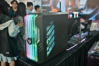 A sleek, modern gaming computer case featuring colorful RGB lighting on its front panel is displayed. The case is placed on a black table alongside several computer monitors. Several people are gathered around, possibly at a tech or gaming event showcase.