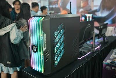 A sleek, modern gaming computer case featuring colorful RGB lighting on its front panel is displayed. The case is placed on a black table alongside several computer monitors. Several people are gathered around, possibly at a tech or gaming event showcase.