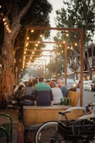 A group of people is gathered around an outdoor seating area under a canopy of string lights. The setting appears to be a street, with trees lining the sidewalk and cars parked along the road. The atmosphere conveys a casual, communal gathering in warm lighting.