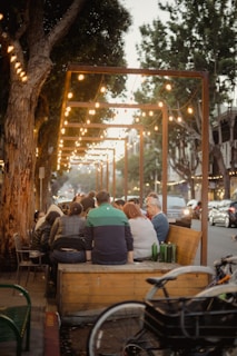 A group of people is gathered around an outdoor seating area under a canopy of string lights. The setting appears to be a street, with trees lining the sidewalk and cars parked along the road. The atmosphere conveys a casual, communal gathering in warm lighting.