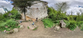 A panoramic view of an outdoor climbing area featuring a large rock face surrounded by lush greenery. Three people are actively engaged in climbing and belaying on the rock surface. Trees and bushes occupy the surrounding landscape, and scattered rocks are visible on the ground.