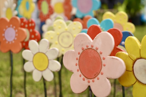 Colorful, decorative flowers are arranged closely in a garden setting. The flowers are crafted from metal or a similar material, each painted in bright hues such as pink, yellow, orange, and blue. The designs feature whimsical patterns with dots and circles at the center. Green grass is visible in the background, enhancing the vibrant and playful atmosphere.