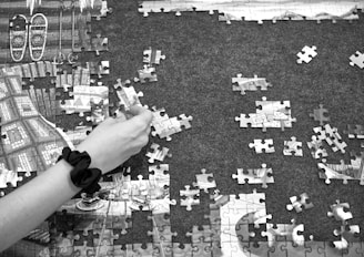 A hand with a black wristband is assembling pieces of a jigsaw puzzle on a dark surface. Some sections of the puzzle are already completed, revealing parts of an image featuring snowshoes and wooden textures. Other puzzle pieces are scattered around, with some turned upside down.