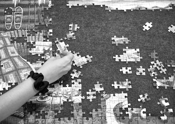 A hand with a black wristband is assembling pieces of a jigsaw puzzle on a dark surface. Some sections of the puzzle are already completed, revealing parts of an image featuring snowshoes and wooden textures. Other puzzle pieces are scattered around, with some turned upside down.