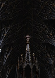 An intricately designed Gothic cathedral ceiling with numerous ribbed arches and pointed details. At the center, a tall ornate structure adorned with gold embellishments supports a purple cross. The patterns and architecture of the ceiling are symmetrical and detailed, creating an impression of depth.