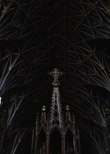 An intricately designed Gothic cathedral ceiling with numerous ribbed arches and pointed details. At the center, a tall ornate structure adorned with gold embellishments supports a purple cross. The patterns and architecture of the ceiling are symmetrical and detailed, creating an impression of depth.