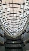 An indoor architectural structure features a large, grid-patterned skylight with curved glass panes. The surrounding walls are smooth, with multiple levels and balconies that curve inward toward the open central space.