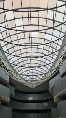 An indoor architectural structure features a large, grid-patterned skylight with curved glass panes. The surrounding walls are smooth, with multiple levels and balconies that curve inward toward the open central space.