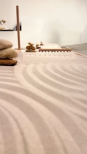 A serene arrangement of stones stacked carefully on a surface, likely part of a Zen garden setup. Smooth sand raked into gentle, flowing patterns surrounds the stones. The soft lighting and minimalist design contribute to a calm and contemplative atmosphere.