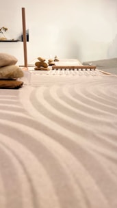 A serene arrangement of stones stacked carefully on a surface, likely part of a Zen garden setup. Smooth sand raked into gentle, flowing patterns surrounds the stones. The soft lighting and minimalist design contribute to a calm and contemplative atmosphere.