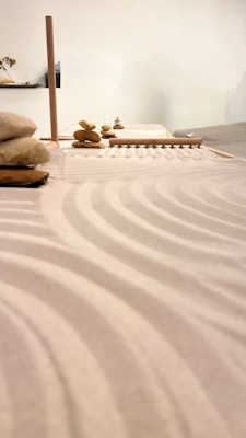 A serene arrangement of stones stacked carefully on a surface, likely part of a Zen garden setup. Smooth sand raked into gentle, flowing patterns surrounds the stones. The soft lighting and minimalist design contribute to a calm and contemplative atmosphere.