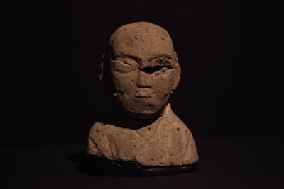 A clay sculpture of a human bust is set against a dark background. The sculpture has a rough texture and portrays a bald figure with minimal facial features. The lighting casts a shadow, enhancing the contours of the face and adding depth to the artwork.