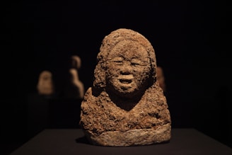 A textured, weathered stone sculpture depicting a human face with closed eyes and a serene expression. The sculpture is prominently placed against a dark background, creating a striking contrast. Two blurred sculptures are visible in the background, adding depth to the composition.