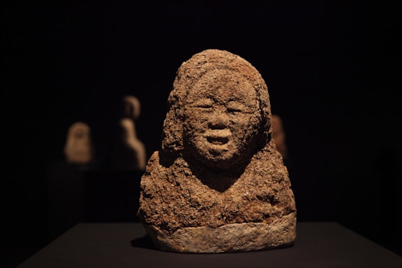 A textured, weathered stone sculpture depicting a human face with closed eyes and a serene expression. The sculpture is prominently placed against a dark background, creating a striking contrast. Two blurred sculptures are visible in the background, adding depth to the composition.