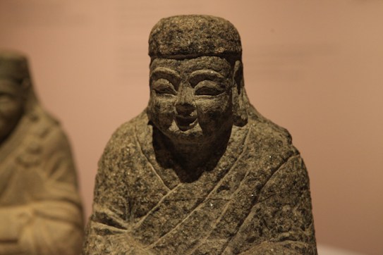 A stone sculpture of a figure wearing robes, featuring a serene expression and detailed facial features. The sculpture is set against a neutral, out-of-focus background, suggesting a museum or gallery setting.