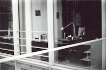 A modern, glass-enclosed office space with a desk visible through the windows. The desk has a lamp, several books or documents, an anglepoise lamp, a computer monitor, and some cables. There are shelves with more books and another desk further inside the room. The image is in black and white.
