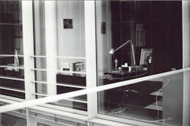 A modern, glass-enclosed office space with a desk visible through the windows. The desk has a lamp, several books or documents, an anglepoise lamp, a computer monitor, and some cables. There are shelves with more books and another desk further inside the room. The image is in black and white.