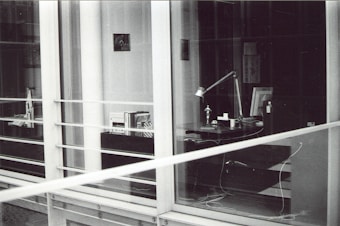 A modern, glass-enclosed office space with a desk visible through the windows. The desk has a lamp, several books or documents, an anglepoise lamp, a computer monitor, and some cables. There are shelves with more books and another desk further inside the room. The image is in black and white.