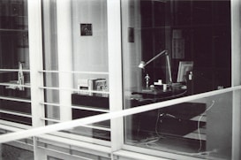 A modern, glass-enclosed office space with a desk visible through the windows. The desk has a lamp, several books or documents, an anglepoise lamp, a computer monitor, and some cables. There are shelves with more books and another desk further inside the room. The image is in black and white.
