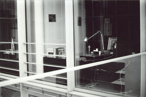 A modern, glass-enclosed office space with a desk visible through the windows. The desk has a lamp, several books or documents, an anglepoise lamp, a computer monitor, and some cables. There are shelves with more books and another desk further inside the room. The image is in black and white.