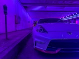 A sleek white sports car is parked in a dimly lit urban setting, illuminated by vibrant purple and blue neon lights. The environment suggests a futuristic or cyberpunk-inspired mood, with the car prominently positioned in the foreground and parking meters along the sidewalk.