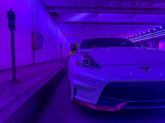 A sleek white sports car is parked in a dimly lit urban setting, illuminated by vibrant purple and blue neon lights. The environment suggests a futuristic or cyberpunk-inspired mood, with the car prominently positioned in the foreground and parking meters along the sidewalk.