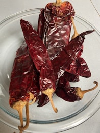 A glass bowl containing several dried red chili peppers with wrinkled, glossy skin and long, curved stems.