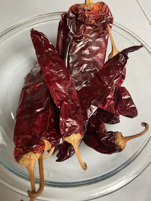 A glass bowl containing several dried red chili peppers with wrinkled, glossy skin and long, curved stems.