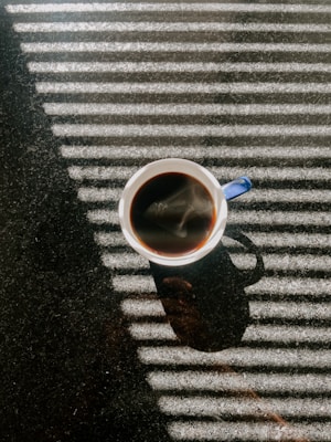 A cup filled with dark coffee is placed on a textured surface, with sunlight streaming through blinds creating a pattern of light and shadow across the scene. The steam rises from the coffee, adding a sense of warmth.