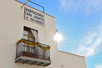 A building with a window featuring a small decorative balcony adorned with tinsel. There is a sign displaying 'Habitaciones Casa Juanita' along with a phone number. The sun is bright, creating a radiant effect in a clear blue sky.