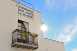 A building with a window featuring a small decorative balcony adorned with tinsel. There is a sign displaying 'Habitaciones Casa Juanita' along with a phone number. The sun is bright, creating a radiant effect in a clear blue sky.