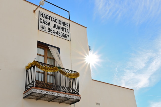 A building with a window featuring a small decorative balcony adorned with tinsel. There is a sign displaying 'Habitaciones Casa Juanita' along with a phone number. The sun is bright, creating a radiant effect in a clear blue sky.