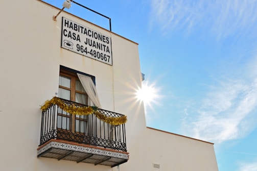 A building with a window featuring a small decorative balcony adorned with tinsel. There is a sign displaying 'Habitaciones Casa Juanita' along with a phone number. The sun is bright, creating a radiant effect in a clear blue sky.