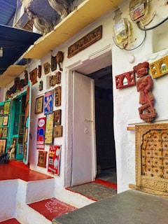 A collection of various vintage and traditional art pieces and decor adorns the exterior wall of a building. The wall includes framed artworks, relief sculptures, and ornate wooden carvings. A wooden statue is prominently displayed next to the doorway, which is slightly ajar. The floor features vibrant red and white patterns, and the overall atmosphere suggests an eclectic and cultural theme.