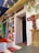 A collection of various vintage and traditional art pieces and decor adorns the exterior wall of a building. The wall includes framed artworks, relief sculptures, and ornate wooden carvings. A wooden statue is prominently displayed next to the doorway, which is slightly ajar. The floor features vibrant red and white patterns, and the overall atmosphere suggests an eclectic and cultural theme.