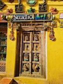 An intricately carved wooden door is surrounded by a bright yellow wall. Above the door is a sign that reads 'Studio art' in colorful letters. Various artistic masks, sculptures, and decor pieces adorn the wall around the door, including depictions of human faces and animals. The door itself features detailed carvings of deities and decorative motifs.