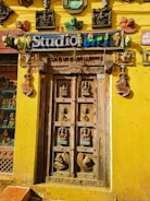 An intricately carved wooden door is surrounded by a bright yellow wall. Above the door is a sign that reads 'Studio art' in colorful letters. Various artistic masks, sculptures, and decor pieces adorn the wall around the door, including depictions of human faces and animals. The door itself features detailed carvings of deities and decorative motifs.