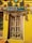 An intricately carved wooden door is surrounded by a bright yellow wall. Above the door is a sign that reads 'Studio art' in colorful letters. Various artistic masks, sculptures, and decor pieces adorn the wall around the door, including depictions of human faces and animals. The door itself features detailed carvings of deities and decorative motifs.