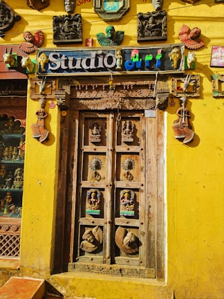 An intricately carved wooden door is surrounded by a bright yellow wall. Above the door is a sign that reads 'Studio art' in colorful letters. Various artistic masks, sculptures, and decor pieces adorn the wall around the door, including depictions of human faces and animals. The door itself features detailed carvings of deities and decorative motifs.