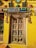 An intricately carved wooden door is surrounded by a bright yellow wall. Above the door is a sign that reads 'Studio art' in colorful letters. Various artistic masks, sculptures, and decor pieces adorn the wall around the door, including depictions of human faces and animals. The door itself features detailed carvings of deities and decorative motifs.