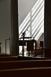 A dimly lit interior with a central podium featuring a microphone and a small floral arrangement. Sunlight streams through slanted blinds, creating striped patterns on the walls and floors. In the background, several empty wooden chairs are partially visible.