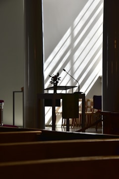 A dimly lit interior with a central podium featuring a microphone and a small floral arrangement. Sunlight streams through slanted blinds, creating striped patterns on the walls and floors. In the background, several empty wooden chairs are partially visible.