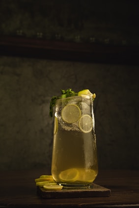 A chilled glass of bright yellow lemonade garnished with fresh mint leaves and lemon slices, set against a crisp white background with lemon and lime greens.