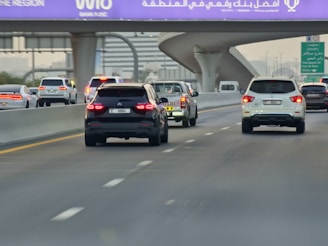 A multi-lane highway with several cars driving in both directions. The vehicles include SUVs, sedans, and a pickup truck. An overhead purple sign with white text is visible above the road, indicating directions or a location. The scene is set during daylight, and the highway is surrounded by urban infrastructure.