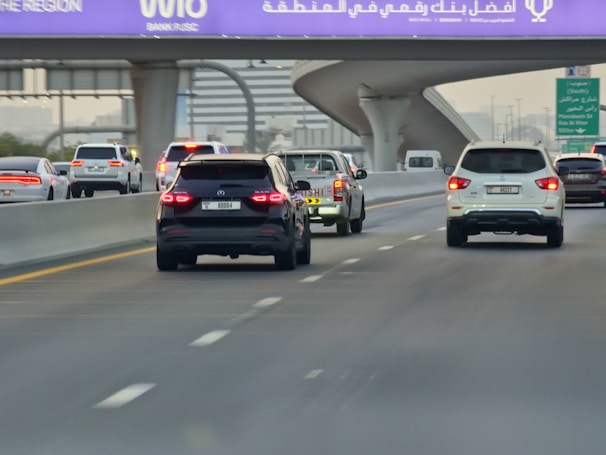 A multi-lane highway with several cars driving in both directions. The vehicles include SUVs, sedans, and a pickup truck. An overhead purple sign with white text is visible above the road, indicating directions or a location. The scene is set during daylight, and the highway is surrounded by urban infrastructure.